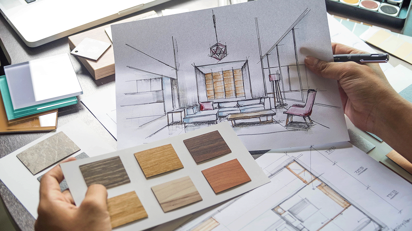 Hands working on an interior design sketch of a furnished room, surrounded by swatches and color palettes