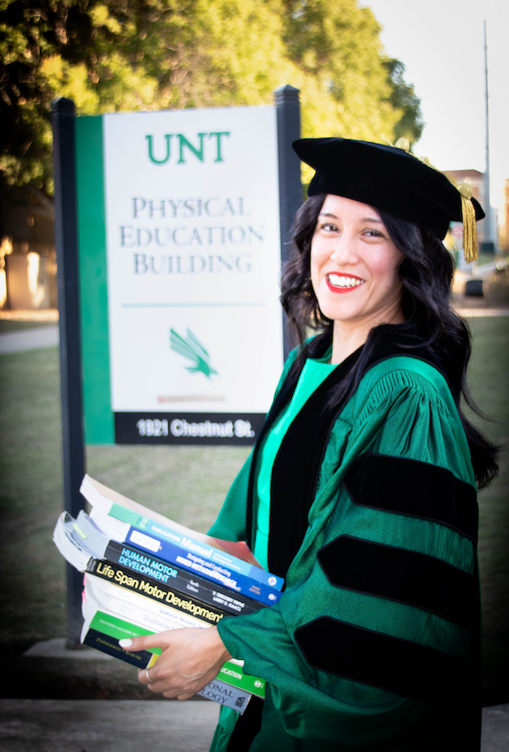 Ana West wearing commencement regalia and holding a stack of books outside the Physical Education building