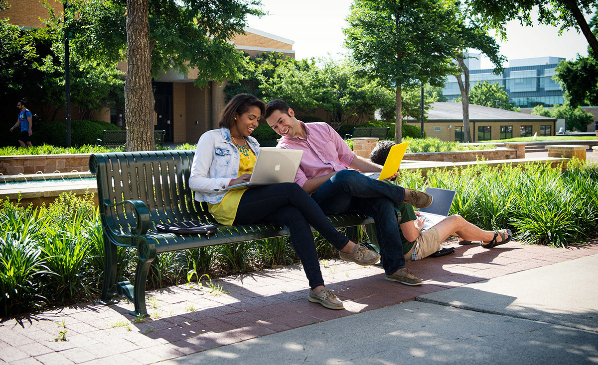 两名学生坐在外面的长凳上，看着笔记本电脑。